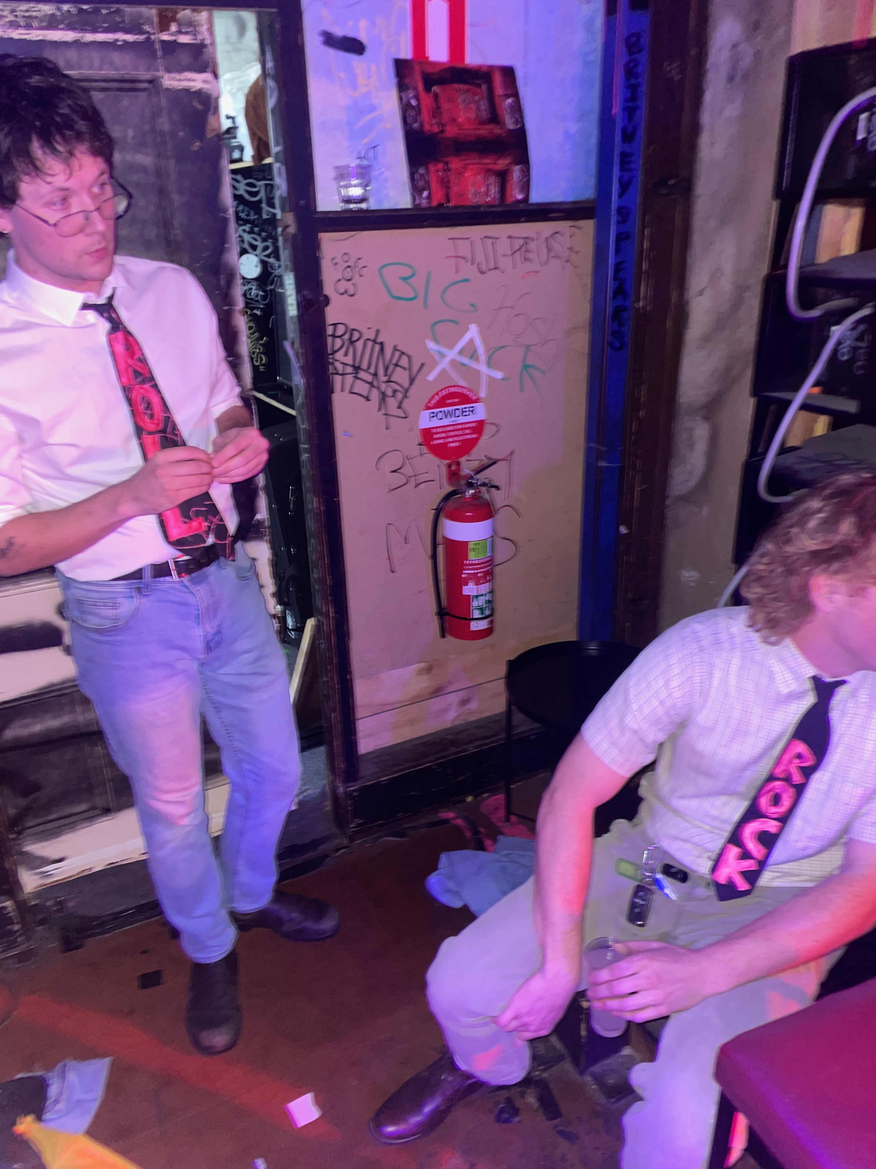Photo of Tom and Tom (our band) wearing white collared shirts and ties with pink block letters sewn onto them, one that says ROCK and one that says ROLL. They are on the corner of the stage at The Tote bandroom.