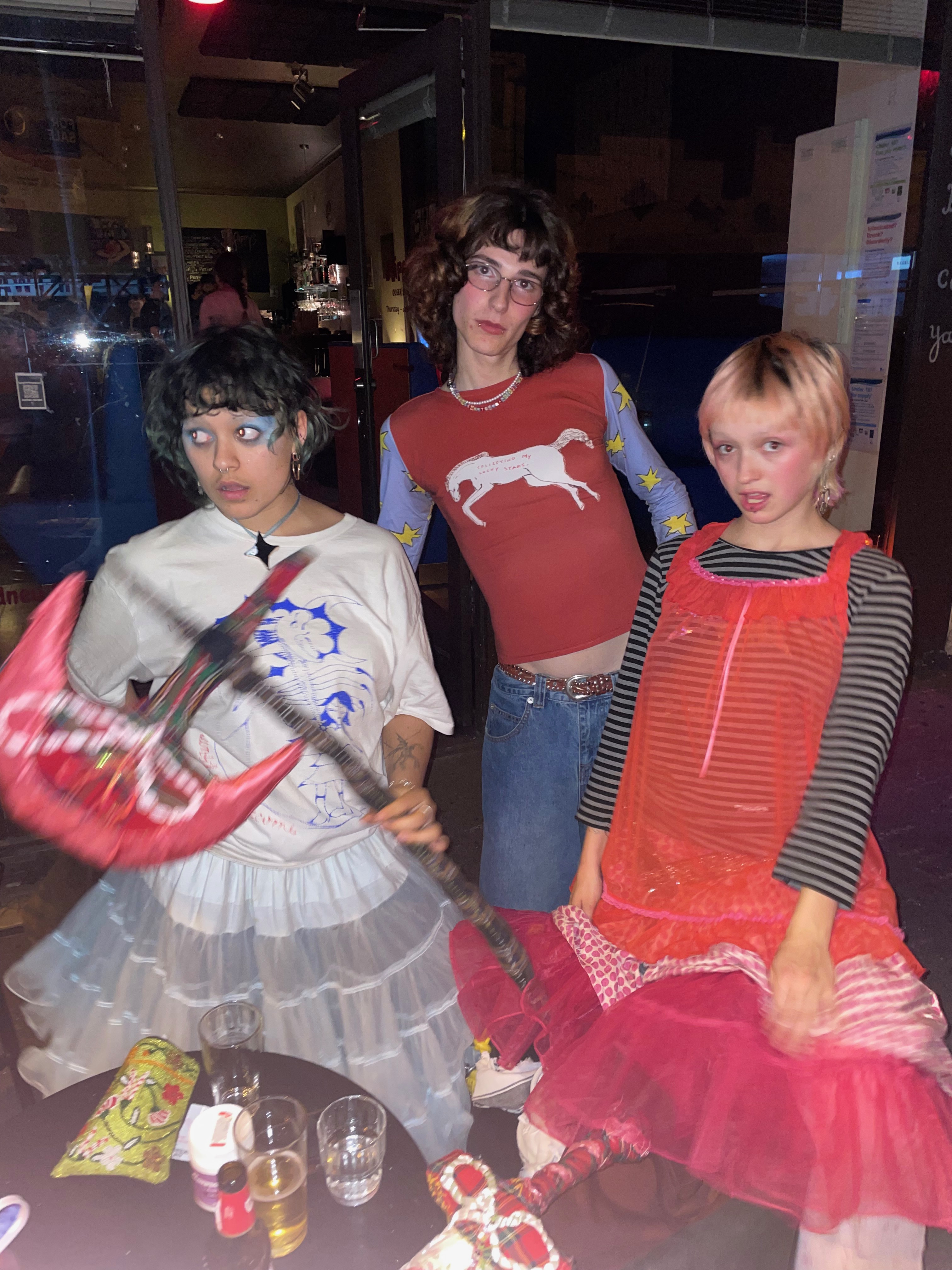 A flash photo of Florence, Lassie and Ada standing out the front of Flippy's Bar. Florence is wearing a blue tutu and a white t-shirt and holding a pink, red and brown large soft sculpture axe. Lassie is wearing a red shirt with a white horse on it. The shirt has longsleeve blue arms with yellow stars, she is also wearing glasses and blue jeans. Ada is wearing a red slip dress with a grey and black long sleeve shirt and a big pink tutu underneath. There is a table in the foreground with some drinks, beer bottles, a small green beaded bag and a soft sculpture sword (only the hilt is visible).
