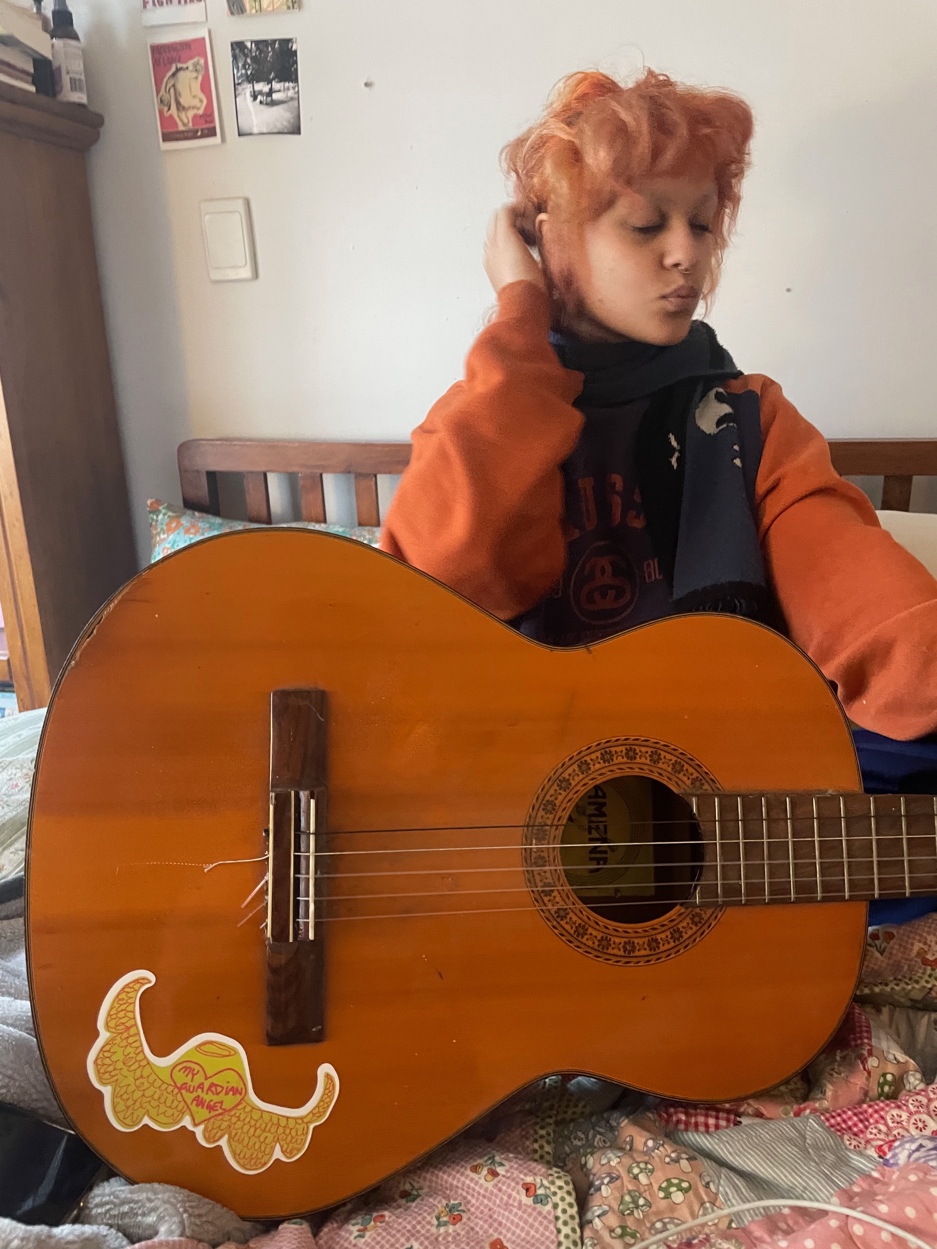 Florence sitting in bed with an acoustic guitar with the My Guardian Angel sticker stuck on it.