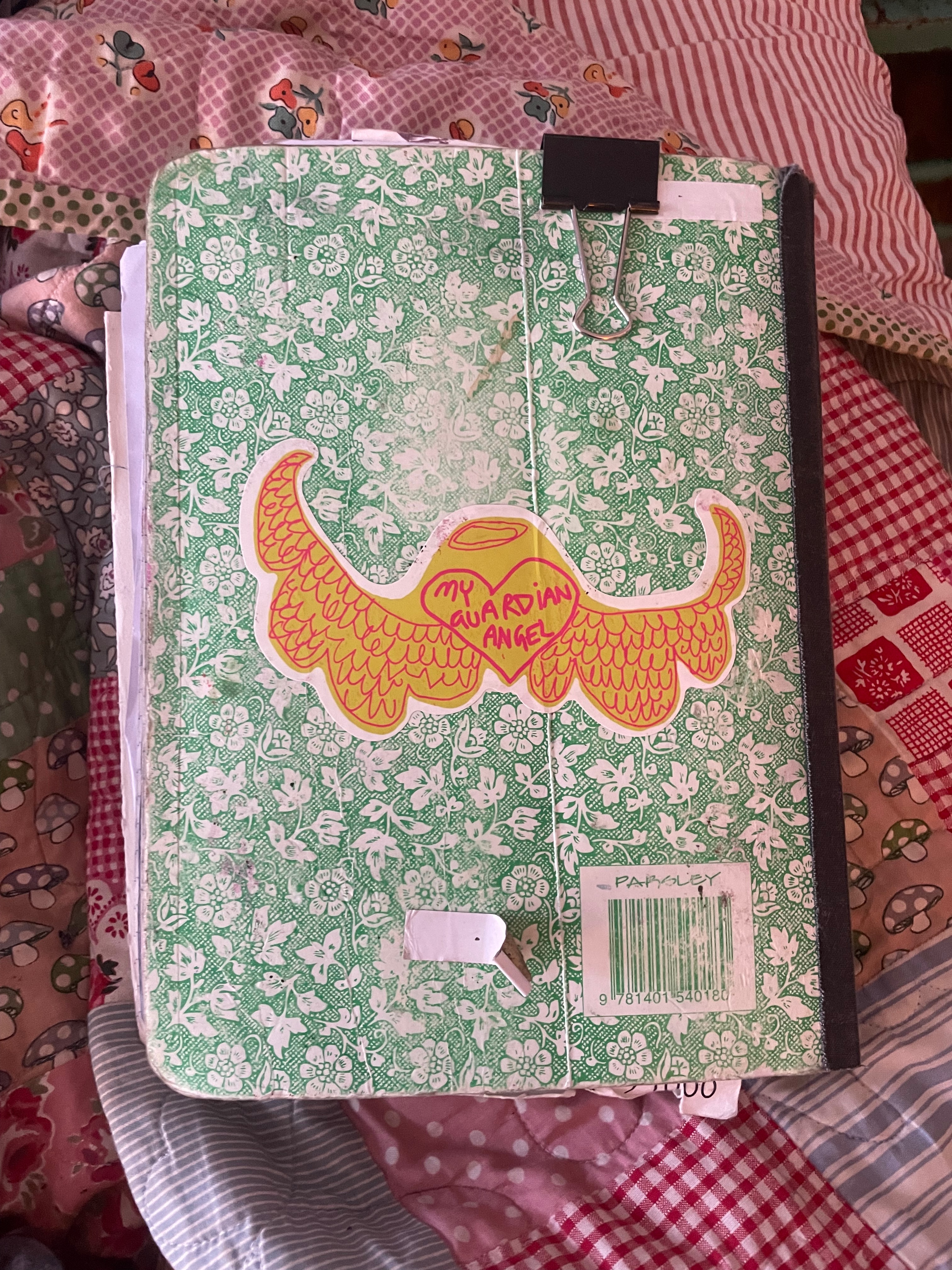 picture of the back side of a green and white floral patterned notebook on top of a pink patchwork blanket. The notebook has a black bulldog clip in the top right of it and a My Guardian Angel sticker of the middle of the cover. The My Guardian Angel sticker is a line drawing of a heart with feathered wings and a halo. Inside the heart it says My Guardian Angel. The linework is neon pink and the background is bright yellow. The sticker is kiss cut to the shape of the winged heart.