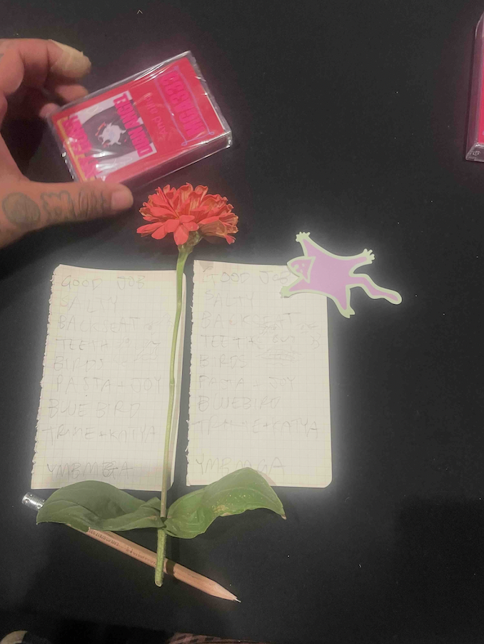 photo of two paper setlists, a drawing by Enoki, an orange zinnea flower, a cassette tape someone is holding